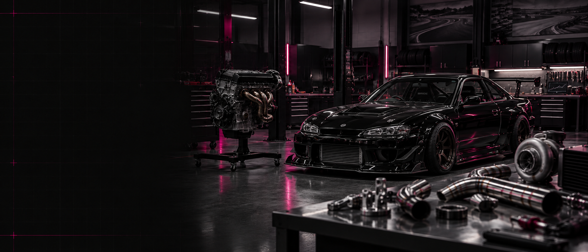 Black performance coupe beside an engine on a stand and fabricated components in a workshop.